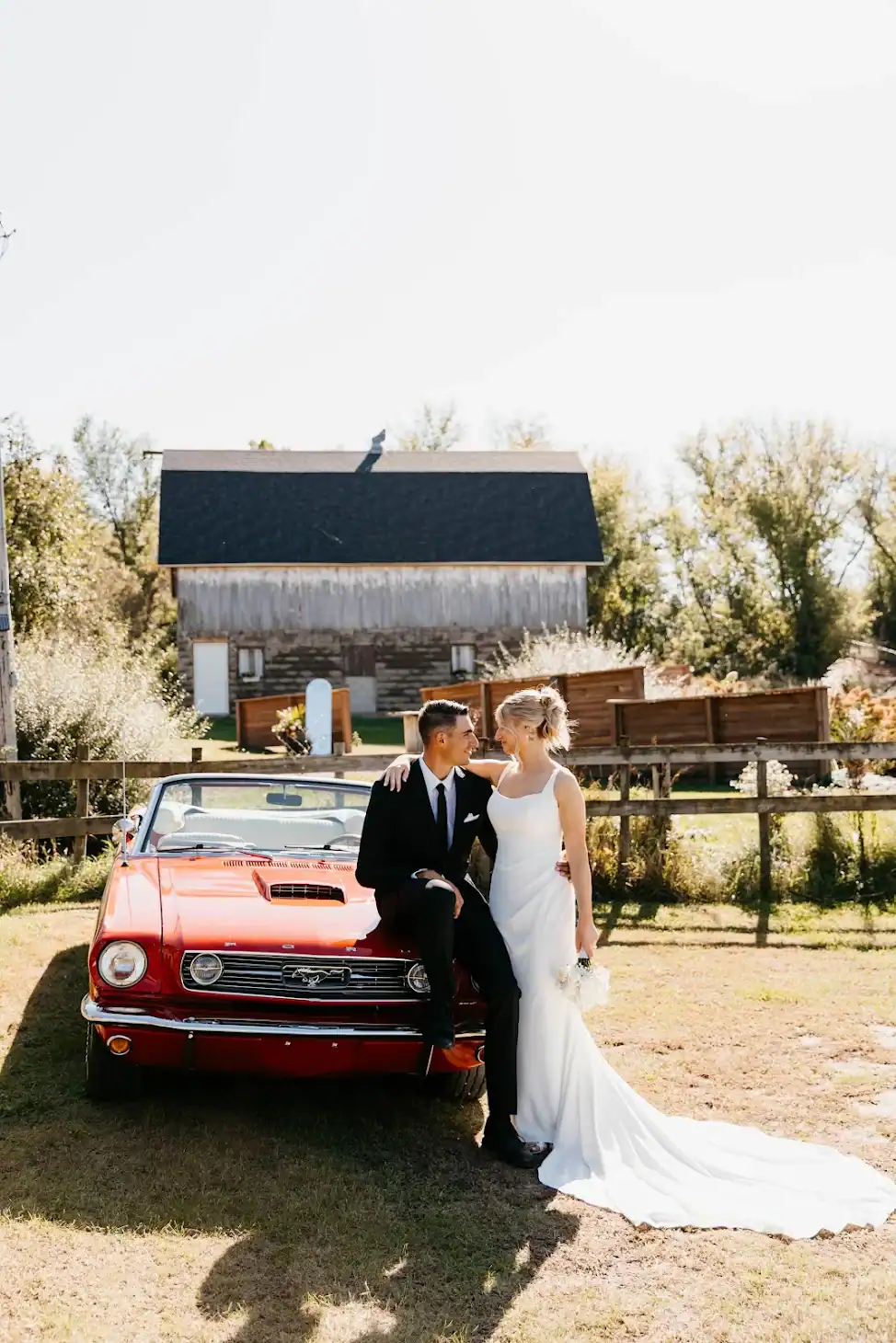 A bride and groom pose beside a red vintage car near a rustic barn under sunny skies on their wedding day.