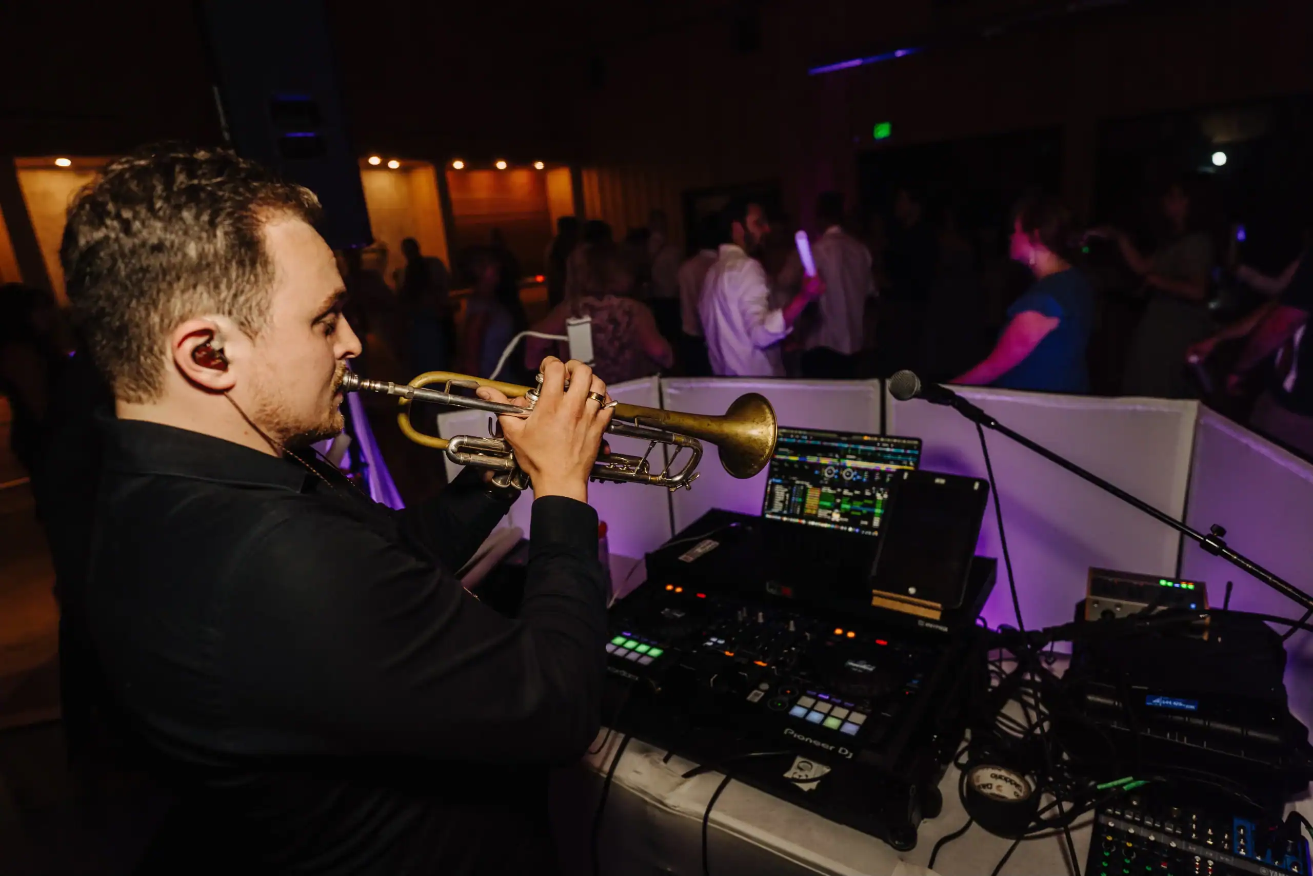 A man plays trumpet next to DJ gear at a dimly lit wedding DJ celebration in Minnesota or Wisconsin.