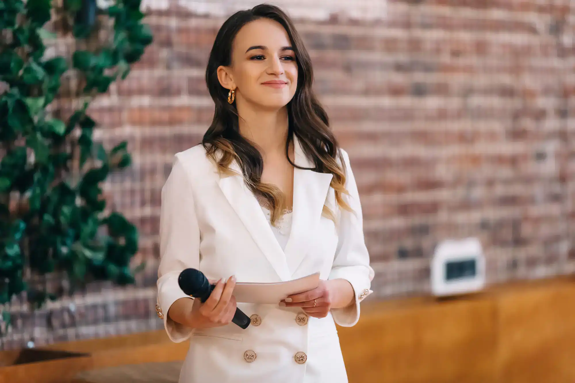 A woman in a white blazer smiles while holding a mic and paper, providing MC services indoors.