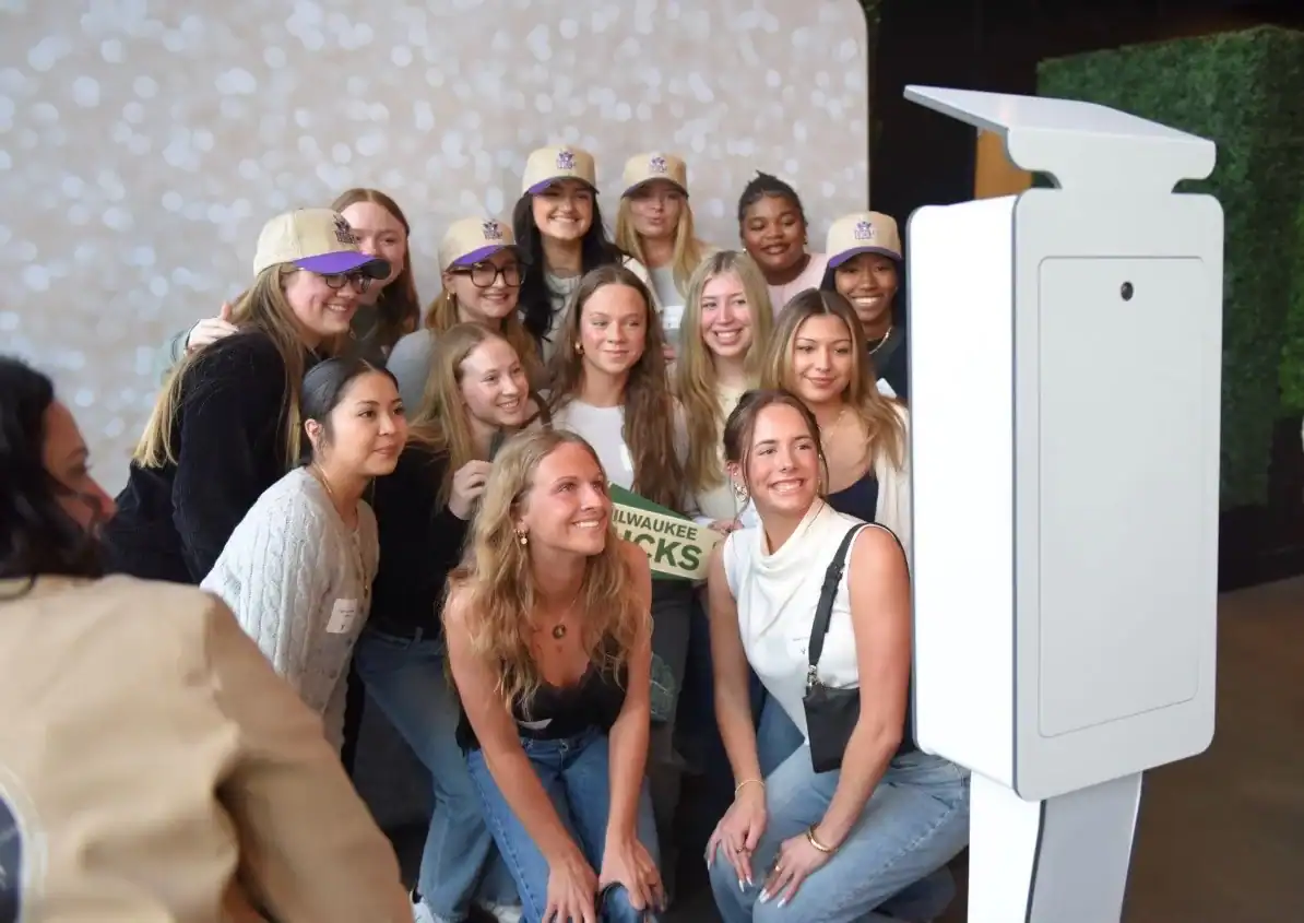 A group of women poses and smiles for a photo in front of a white photo booth at an indoor event.