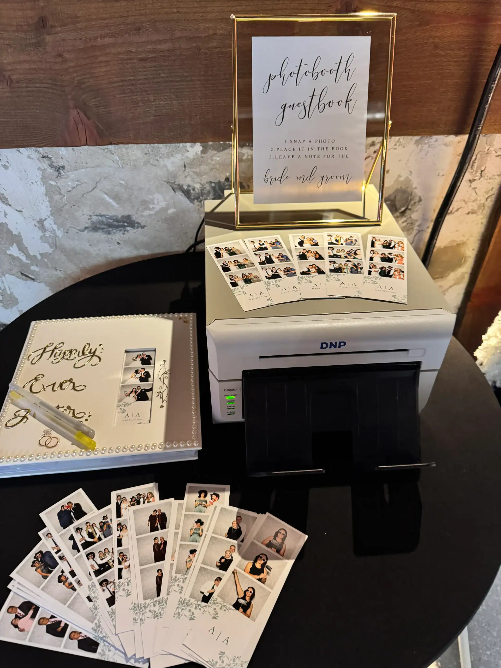 Photo booth guestbook station features photo strips, an album, and instruction sign on a black table.