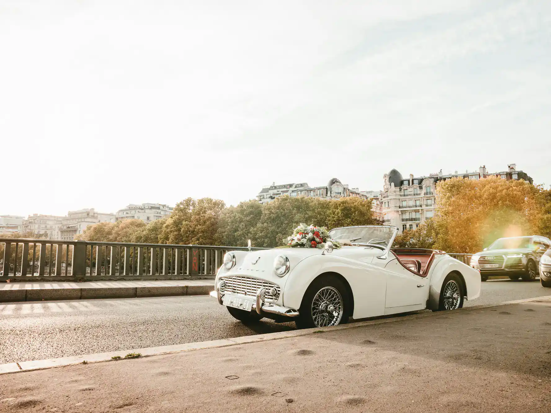 A white vintage convertible decorated with flowers is parked by a railing on a sunny city street.