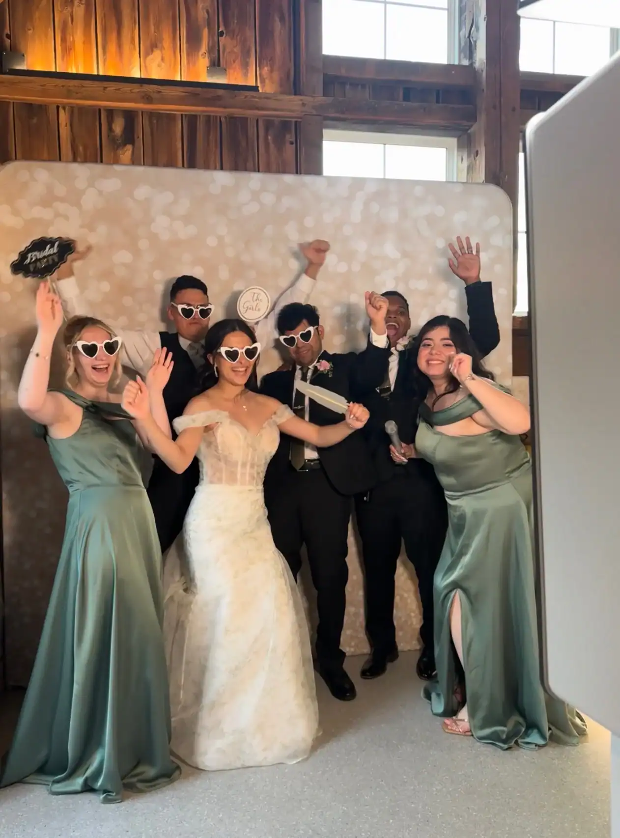 Six people in formal wear pose playfully in a wedding photo booth, all wearing heart-shaped sunglasses.